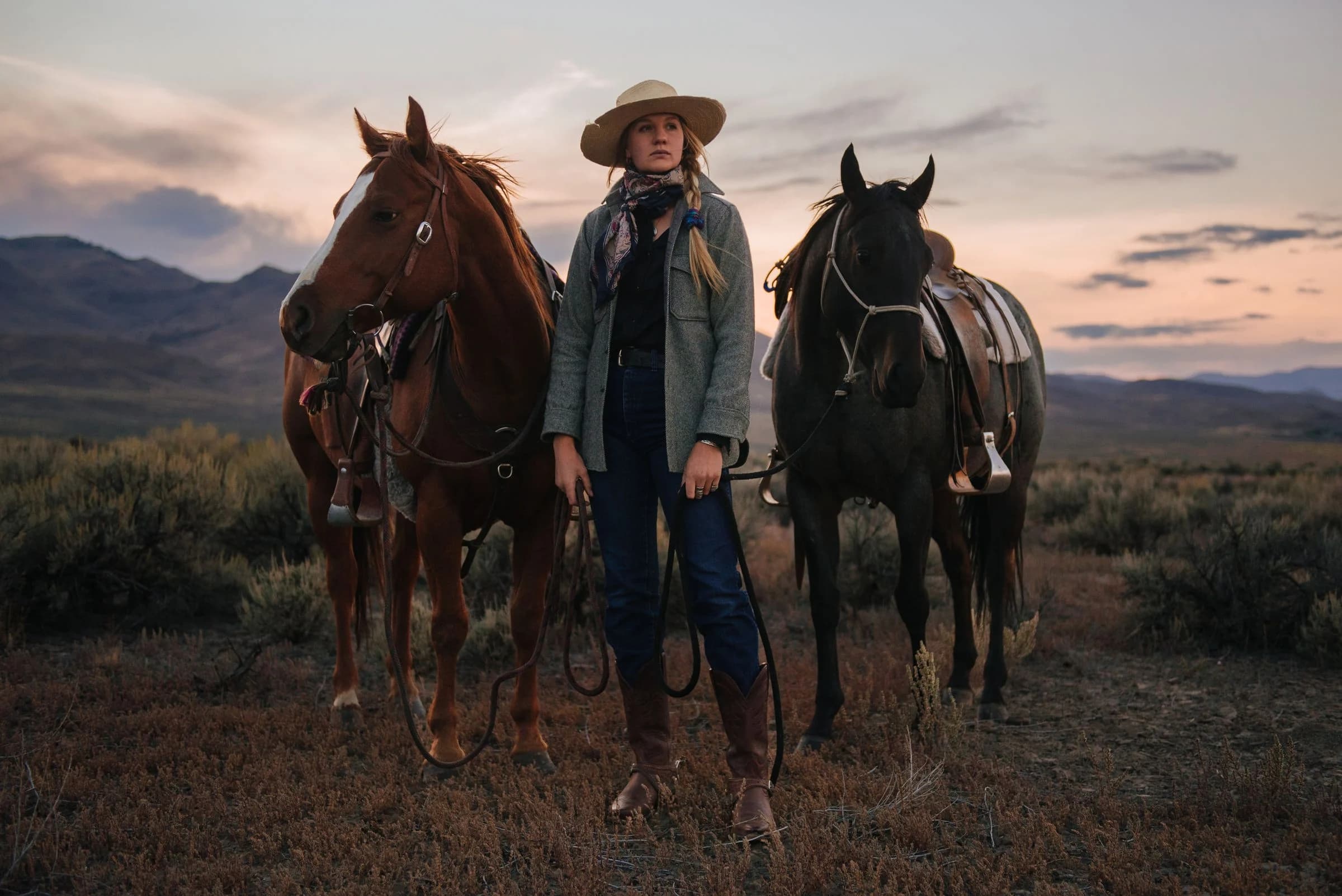 Tecovas ranch photography: a woman in cowboy boots stands between two saddled horses beneath a mountain sunset